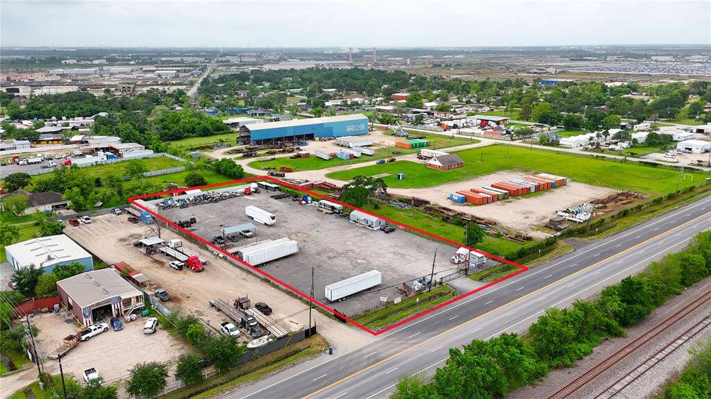 This aerial photo shows a large, fenced commercial lot outlined in red, featuring a paved area with trucks and containers. It's located along a major road, with nearby industrial and green areas, offering easy accessibility and significant space for business operations.