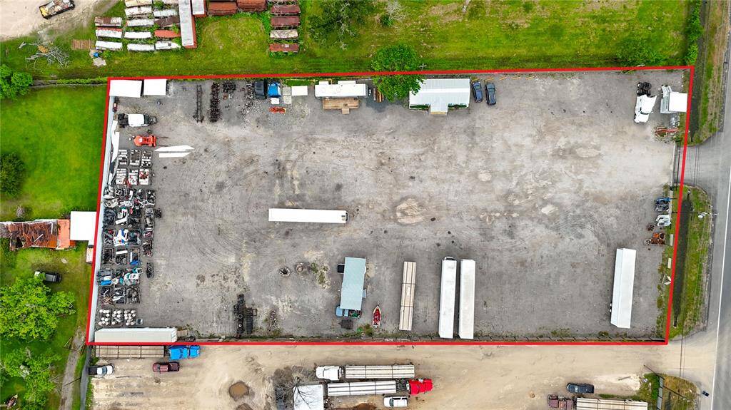 This aerial photo shows a large, fenced commercial lot with a gravel surface, featuring several parked trailers and vehicles. There are small structures along the perimeter, and the lot is surrounded by greenery. Ideal for industrial or storage use.
