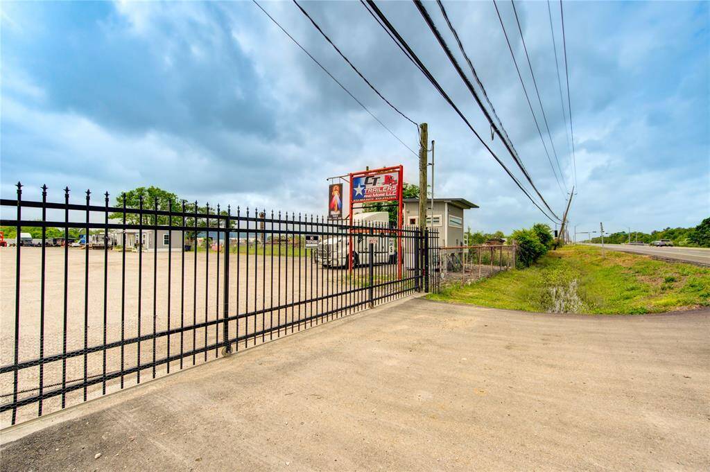 This photo shows a gated commercial lot with a paved entrance. It includes a small building, signage for a trailer business, and a gravel area. The lot is adjacent to a road, offering easy access.