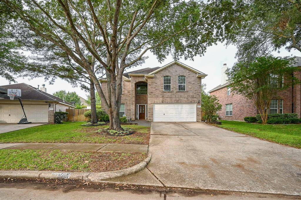 This is a two-story brick home with a spacious driveway and a two-car garage. The front yard features mature trees and well-maintained landscaping, creating an inviting entrance.