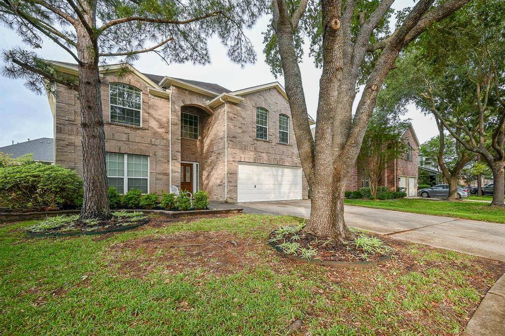 A two-story brick home with a double garage, nestled among mature trees. The well-maintained front yard features lush grass and landscaped garden beds, creating an inviting curb appeal.