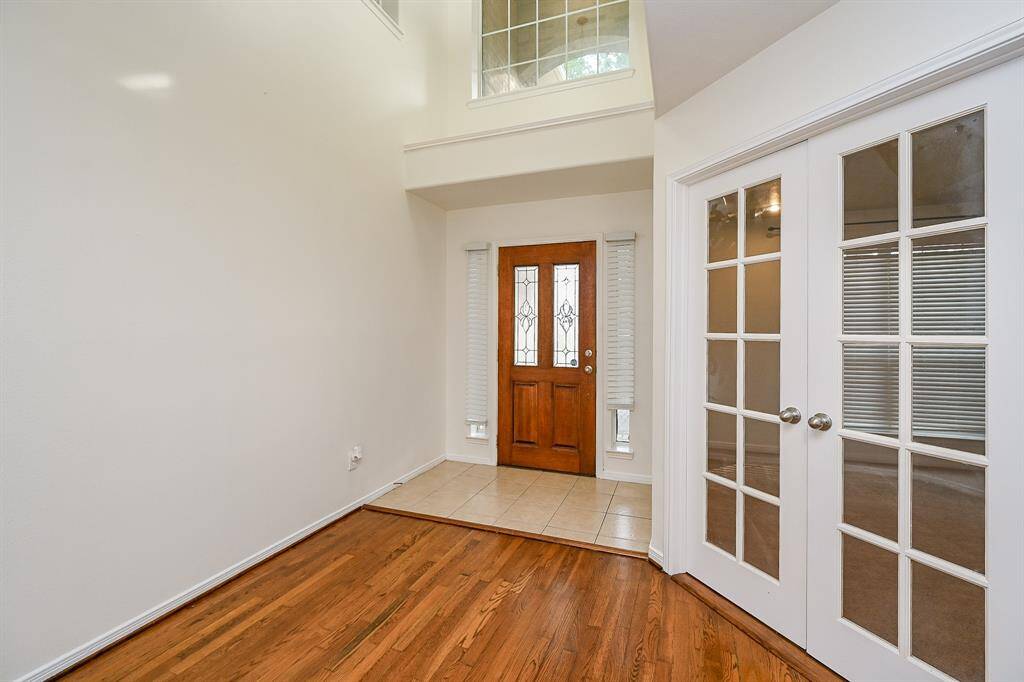 This photo showcases a bright entryway with a high ceiling, wooden flooring, and a wooden front door with decorative glass panels. Adjacent are elegant French doors leading to study, creating an inviting and open atmosphere.