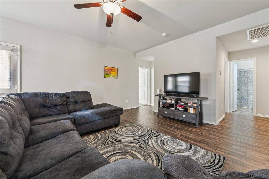 This spacious living room features a cozy sectional sofa, elegant wood flooring, and a ceiling fan. A large TV stands ready for entertainment, and ample natural light streams in from the window, creating a welcoming atmosphere.