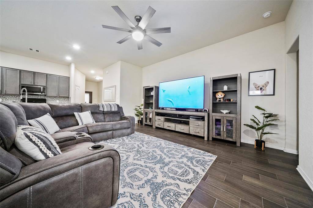 Modern living room with a spacious, open layout featuring ample space for your entertainment center. The area is well-lit with recessed lighting and a ceiling fan. The space includes wood-look tile flooring and provides a seamless transition to the kitchen, creating an inviting atmosphere for relaxation and entertainment.