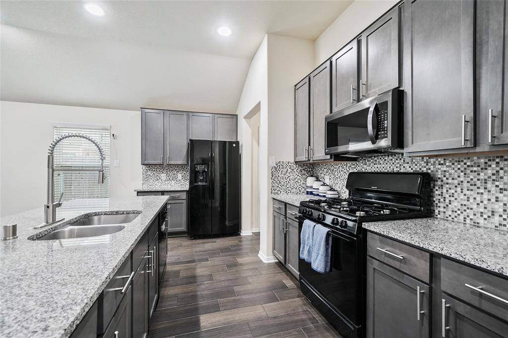 This modern kitchen features sleek gray cabinetry, granite countertops, and a stylish mosaic tile backsplash. It includes stainless steel appliances, a gas stove, and a large double-basin sink with a high-arc faucet. The open layout and recessed lighting enhance the space.
