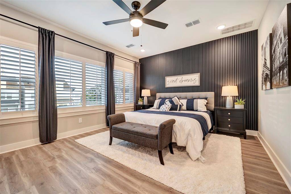 We are invited into the primary suite on the 2nd floor featuring blind, elegant wood plank floors, ceiling fan & beautiful decorative feature wall.