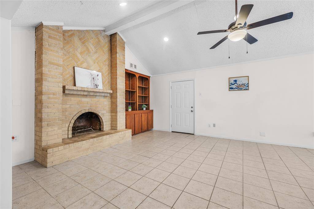 This is a spacious living area featuring a vaulted ceiling with a ceiling fan, tile flooring, a cozy fireplace, and ample natural light from the windows with new 2" blinds.There's also built-in shelving for extra storage or display.