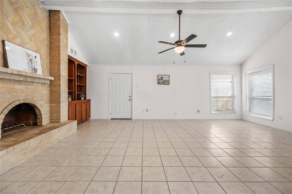 This is a spacious living area featuring a vaulted ceiling with a ceiling fan, tile flooring, a cozy fireplace, and ample natural light from the windows with new 2" blinds.There's also built-in shelving for extra storage or display.