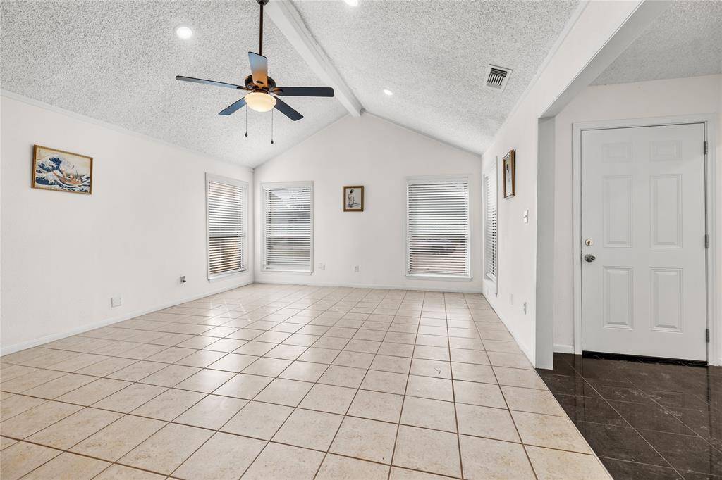 This is a spacious living area featuring a vaulted ceiling with a ceiling fan, tile flooring, a cozy fireplace, and ample natural light from the windows with new 2" blinds.There's also built-in shelving for extra storage or display.