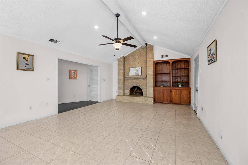This is a spacious living area featuring a vaulted ceiling with a ceiling fan, tile flooring, a cozy fireplace, and ample natural light from the windows with new 2" blinds.There's also built-in shelving for extra storage or display.