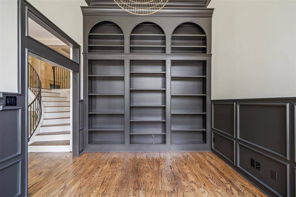 Elegant study features built-in dark wood bookshelves with arch details, complemented by rich wood flooring and wainscoting. A curved staircase with decorative iron railing is visible, adding a touch of sophistication.