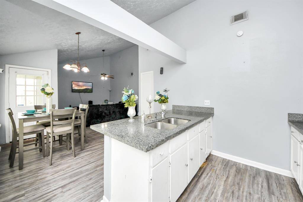 This photo showcases a modern, open-concept kitchen and dining area with gray granite countertops, white cabinetry, and wood-style flooring. The space is well-lit with natural light and features a cozy dining setup adjacent to the living area.