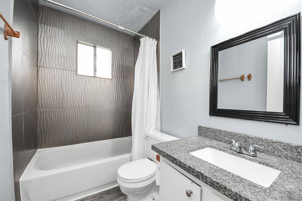 This bathroom features a modern design with textured wall tiles, a bathtub with a shower, and a granite countertop. It includes a large framed mirror and a window for natural light.