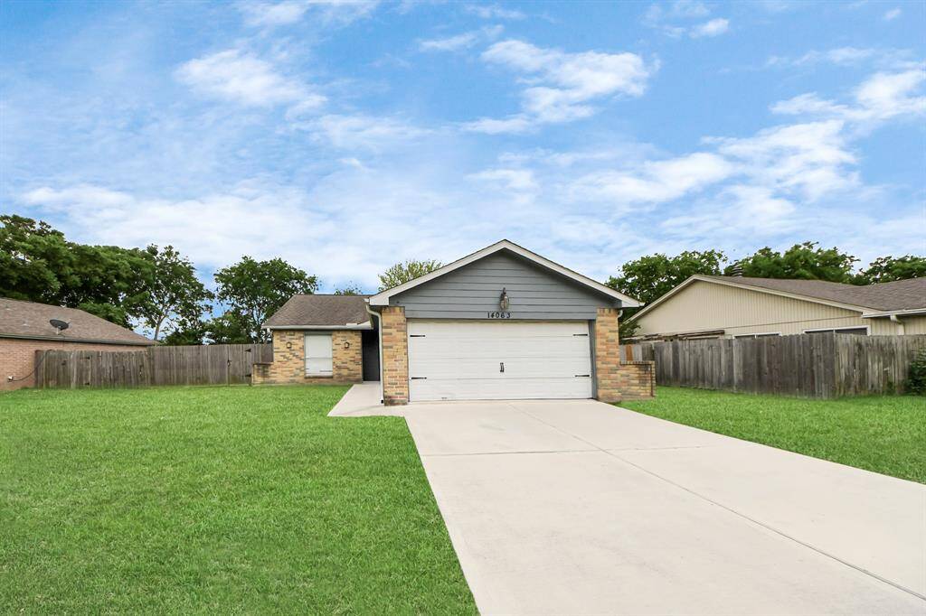 This photo shows a single-story home with a gray and brick exterior, featuring a two-car garage. The house is set back on a spacious green lawn with a long concrete driveway.