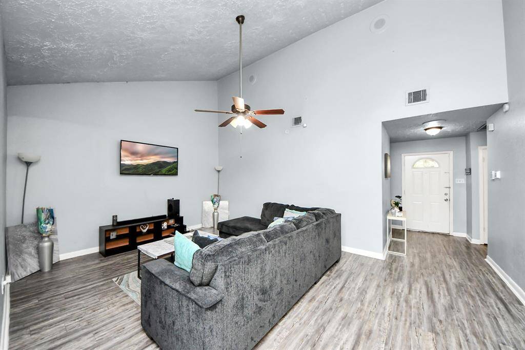 This living room features high ceilings, a modern ceiling fan, and light gray walls and luxury vinyl flooring. The living room is open and inviting, leading to a front entryway with a white door and natural light.