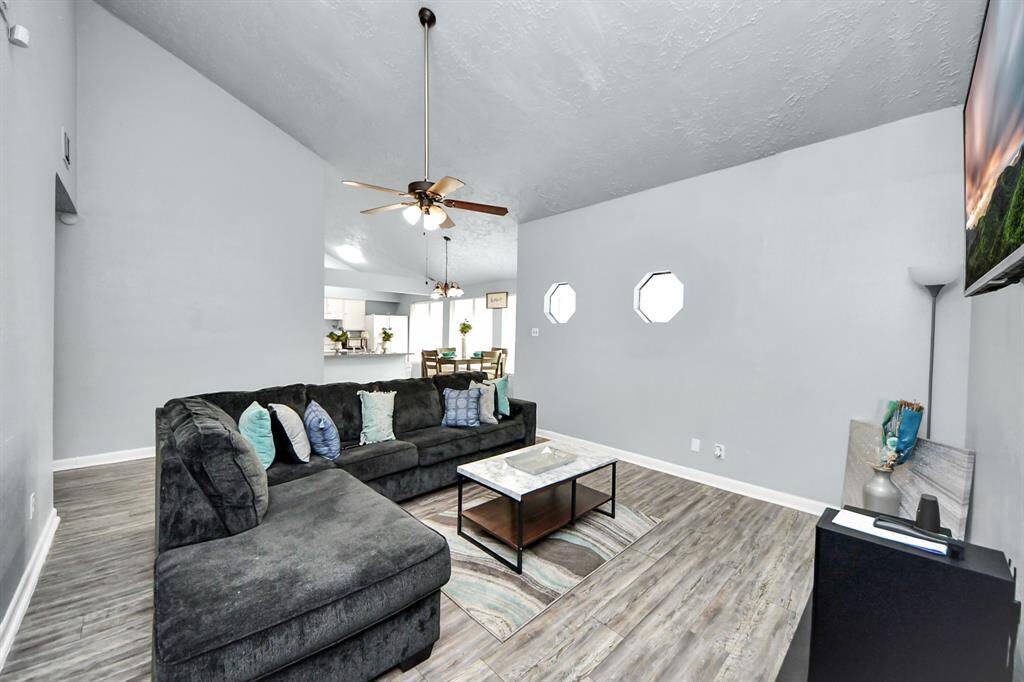 This inviting living room features a vaulted ceiling, modern wood flooring, and ample natural light from octagonal windows. It includes a cozy L-shaped sectional and a ceiling fan, seamlessly connecting to the dining area and kitchen for open-concept living.
