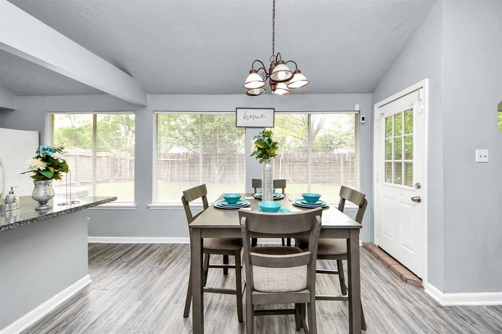Bright dining area with large windows offering plenty of natural light. Features modern gray flooring, a cozy dining set, and a stylish chandelier. The open layout connects to the kitchen, enhancing the space's welcoming feel.
