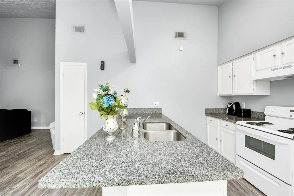 This photo showcases a bright, modern kitchen with granite countertops, white cabinetry, and stainless steel appliances. The open layout and high ceiling create a spacious feel, while the adjacent living area features stylish wood flooring.