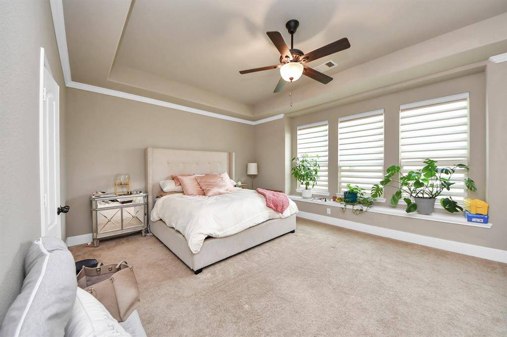 Spacious primary bedroom with neutral tones, featuring a tray ceiling and ceiling fan. Large windows provide plenty of natural light.