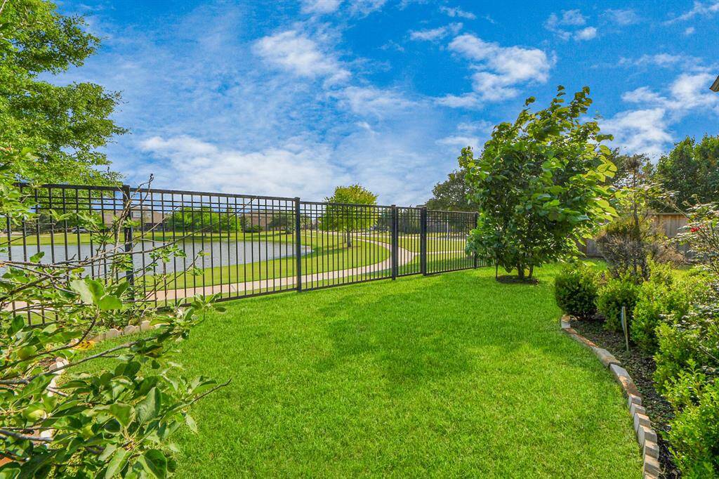 Lush, green backyard with a metal fence overlooking a serene pond and walking path, offering a peaceful and scenic view.