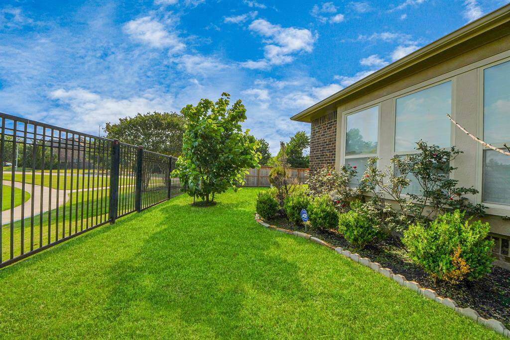 Beautifully maintained backyard featuring lush green grass, a variety of shrubs, and a small tree. It is bordered by a black metal fence.