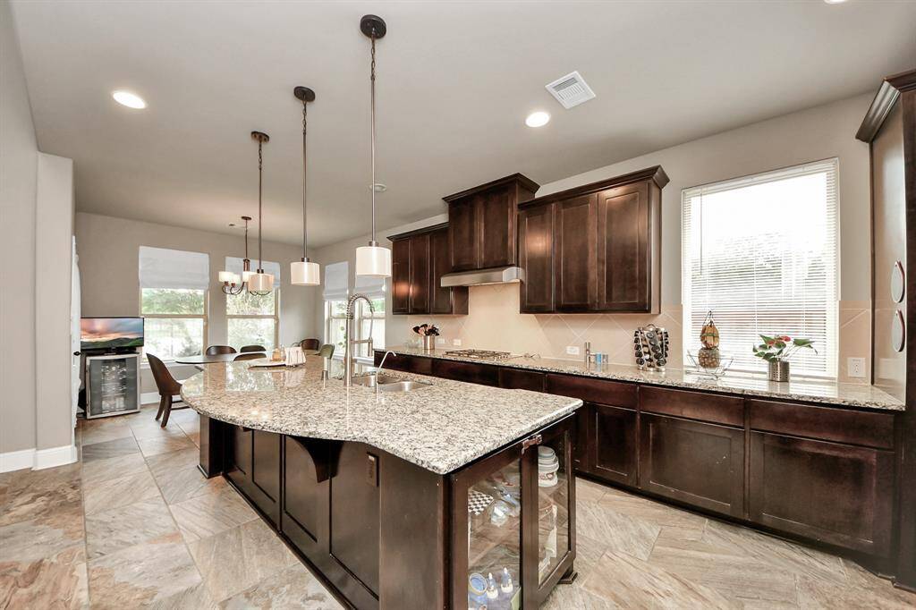 Spacious kitchen with dark wood cabinetry, granite countertops, large central island with sink, and modern pendant lighting. Ample natural light from multiple windows, open layout leading to dining area.