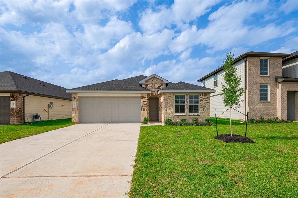 A wide, extended driveway welcomes you home, leading to a two-car garage seamlessly integrated into the classic brick and stone façade. A young tree stands in the lush green lawn, adding a touch of natural charm to the landscape. The recessed front entryway offers both privacy and architectural interest.