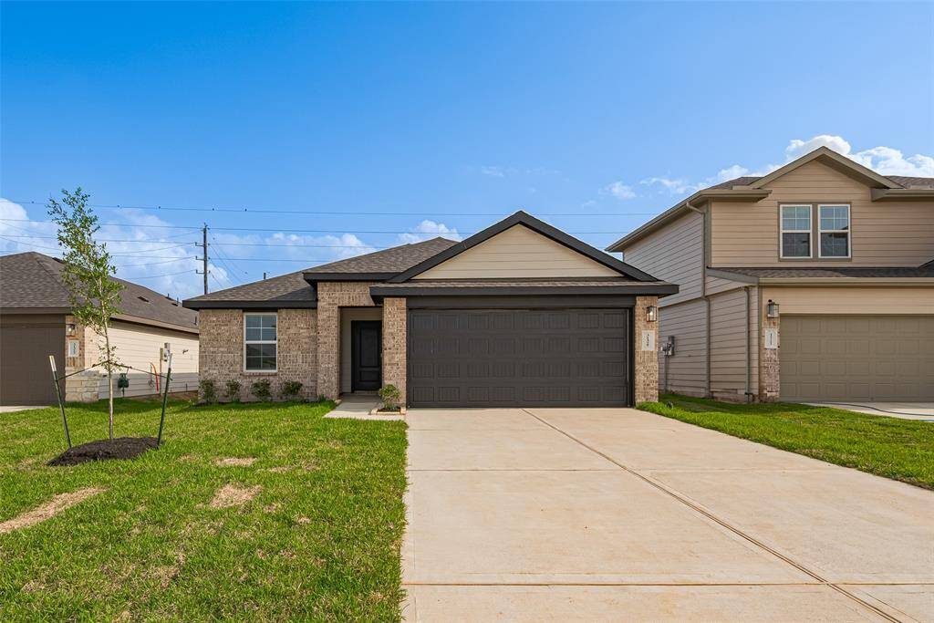 A wide, extended driveway leads to the two-car garage with striking dark-paneled doors that echo the home’s front entry. The brick and light-toned facade is complemented by a manicured lawn that adds a fresh burst of greenery to the curb appeal.