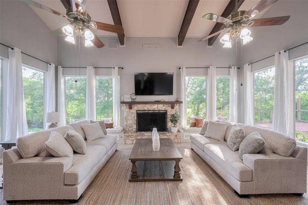 This spacious living room is perfect for relaxation and entertaining, with large windows that let in abundant natural light, offering a serene view of the outdoor greenery. The vaulted ceilings and rustic wooden beams create a grand yet inviting atmosphere, ideal for cozy nights or social gatherings. The stone fireplace adds warmth and charm to this beautiful space.