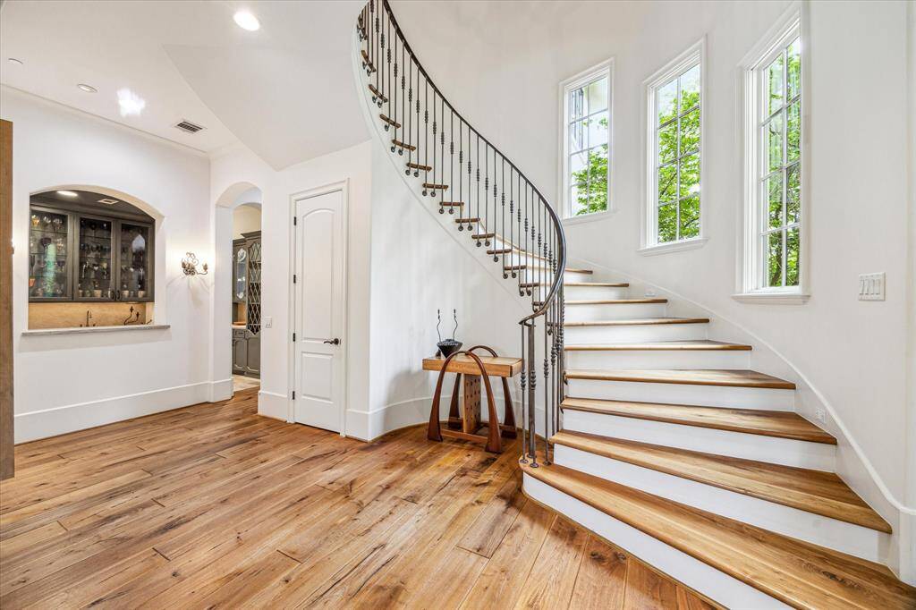 Elegant staircase off the foyer with graceful wrought iron railing, large windows offering serene views of the lush backyard and sparkling pool, complemented by warm wide plank wood floors.