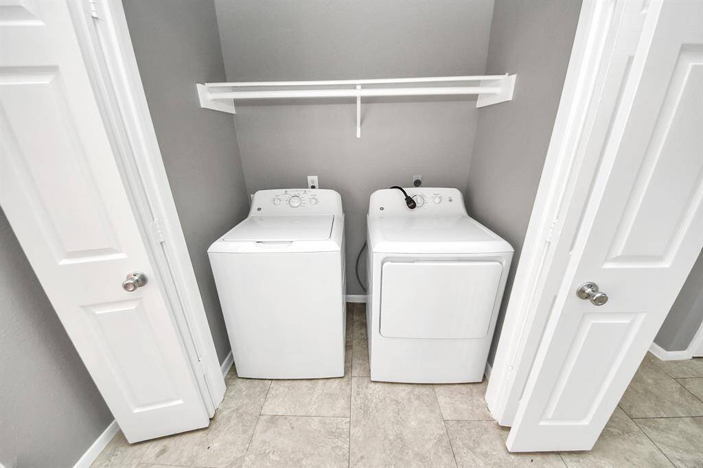 This photo shows a compact laundry nook with a washer and dryer, enclosed by white double doors. It features a shelf above for additional storage and has a clean, modern look with neutral-colored walls and tile flooring.