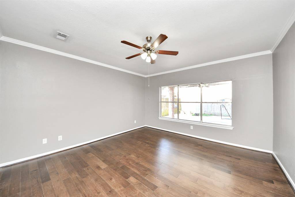 This spacious room features elegant wood flooring, neutral gray walls, and a ceiling fan. A large window offers ample natural light and a view of the backyard.