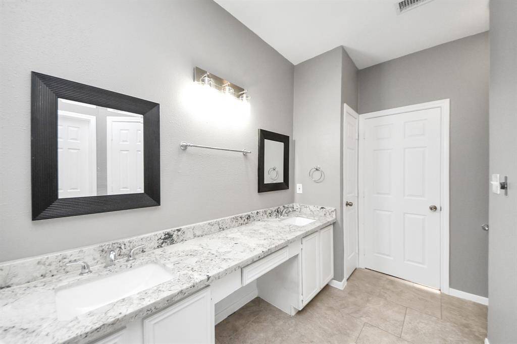 This bathroom features a sleek, modern design with a dual sink vanity and granite countertops. The neutral gray walls and tile flooring create a clean and spacious feel, complemented by framed mirrors and a bright light fixture.