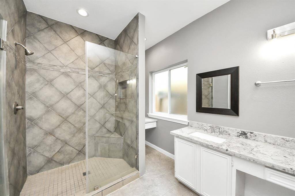 This bathroom features a modern design with a spacious walk-in shower, elegant grey tile walls, and a sleek glass door. The vanity includes a white cabinet with a granite countertop, complemented by a large window for natural light and a stylish framed mirror.