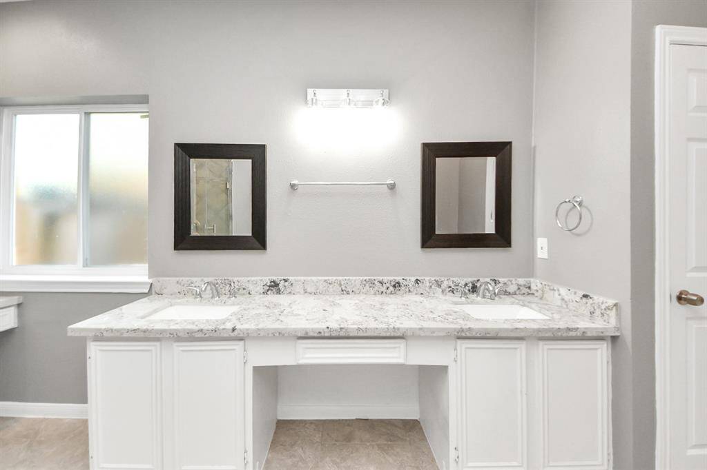 This bathroom features a double vanity with a sleek granite countertop, two framed mirrors, and neutral gray walls. Ample natural light comes through a frosted window, creating a bright and inviting space.