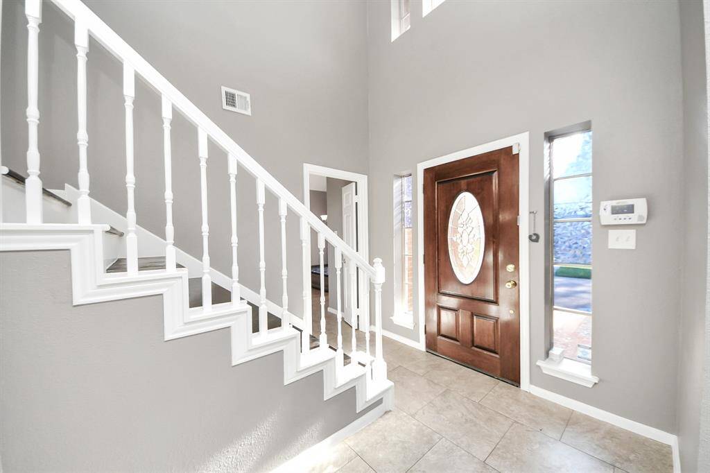 Bright entryway featuring a wooden front door with decorative glass, tiled flooring, and a staircase with white railing. Large windows allow for natural light, creating a welcoming atmosphere.