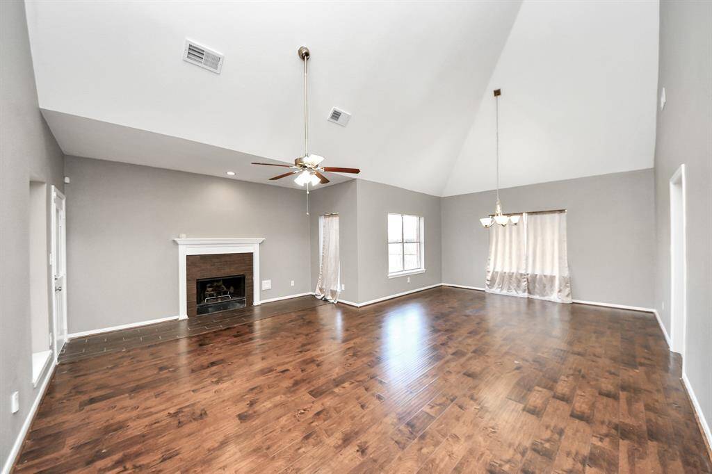 Spacious living room with vaulted ceilings, hardwood floors, and a cozy fireplace. Large windows provide ample natural light, and there's a central ceiling fan and chandelier for added elegance.
