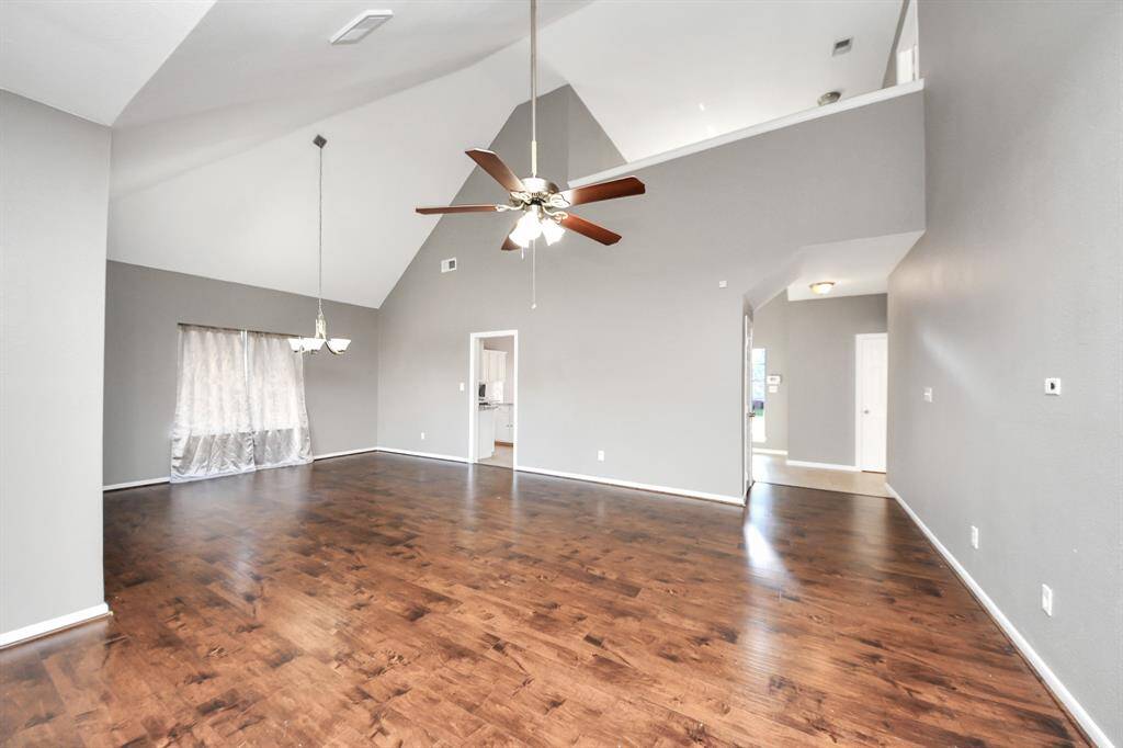 This spacious living room features high vaulted ceilings, hardwood floors, and neutral gray walls. It includes a ceiling fan and a chandelier, with natural light streaming through large windows, creating a bright and open atmosphere.