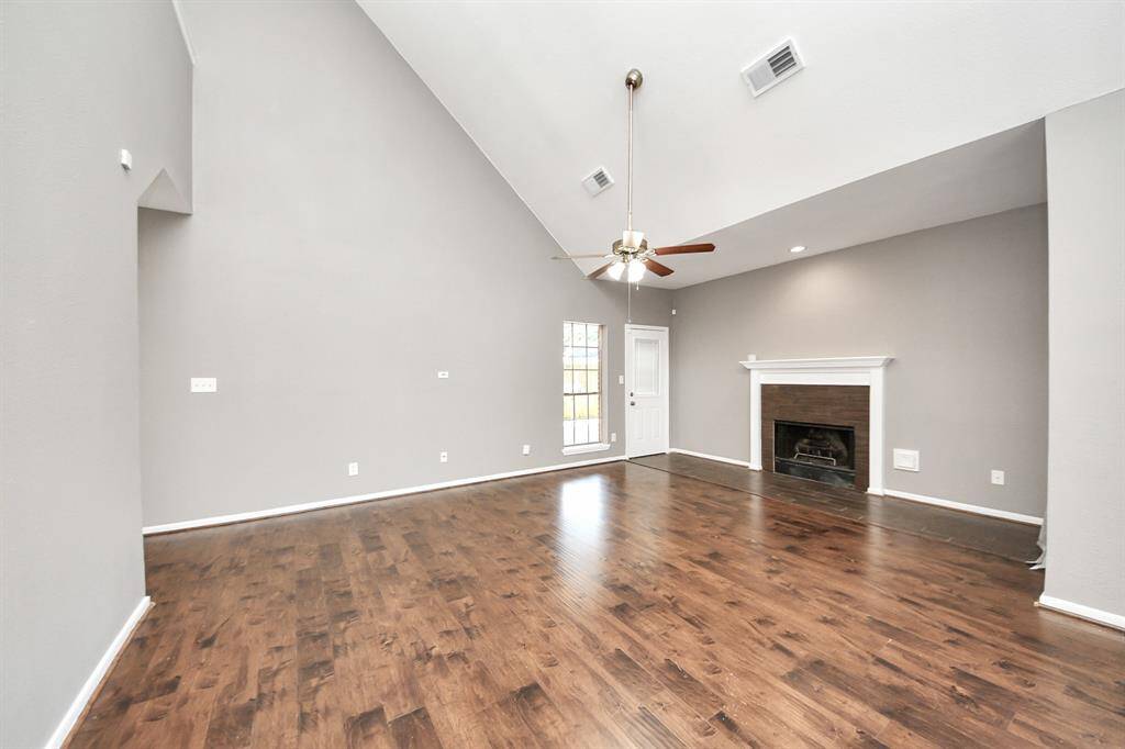 This spacious living room features high vaulted ceilings, a ceiling fan, and a cozy fireplace. The room is accented with light gray walls and rich wooden flooring, creating a warm and inviting atmosphere. Natural light streams in through a door with glass panes, enhancing the open feel.
