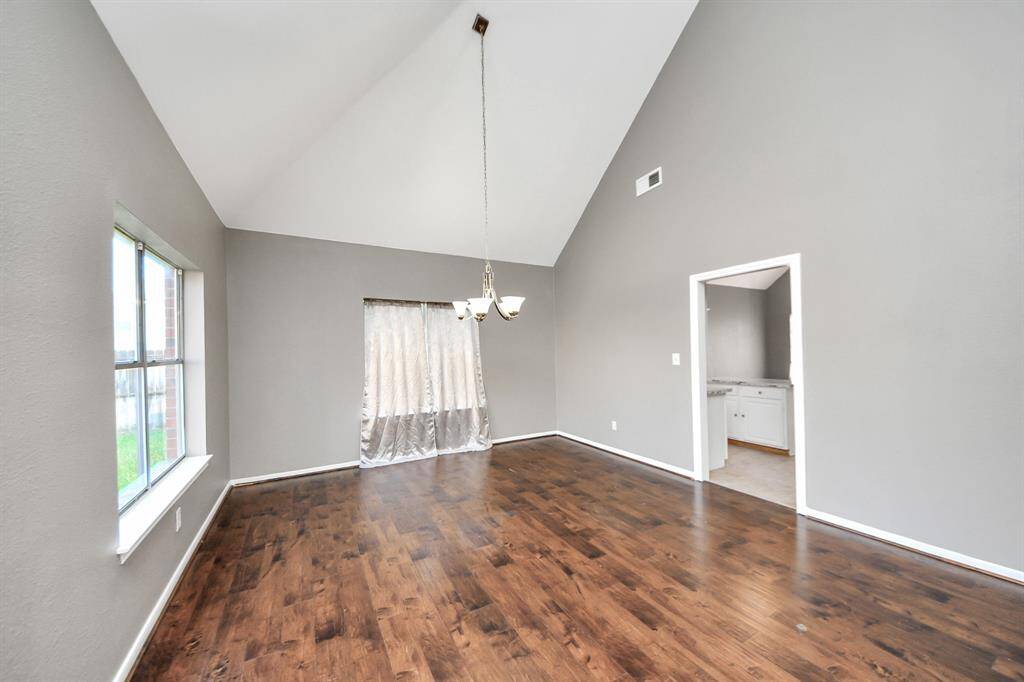 This room features high vaulted ceilings, hardwood floors, and neutral gray walls. It includes a large window for natural light, a hanging light fixture, and an adjoining room with white cabinetry visible through a doorway.
