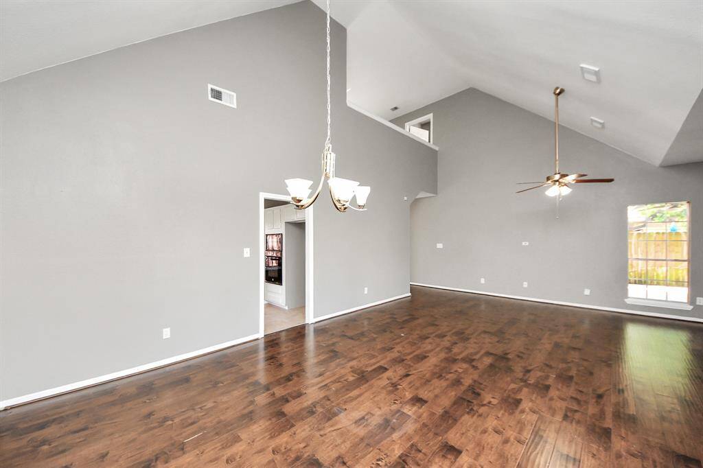This spacious living area features high vaulted ceilings, rich wood flooring, and abundant natural light from large windows. A modern chandelier and ceiling fan add elegance and comfort. The open layout seamlessly connects to adjacent rooms, enhancing flow and functionality.