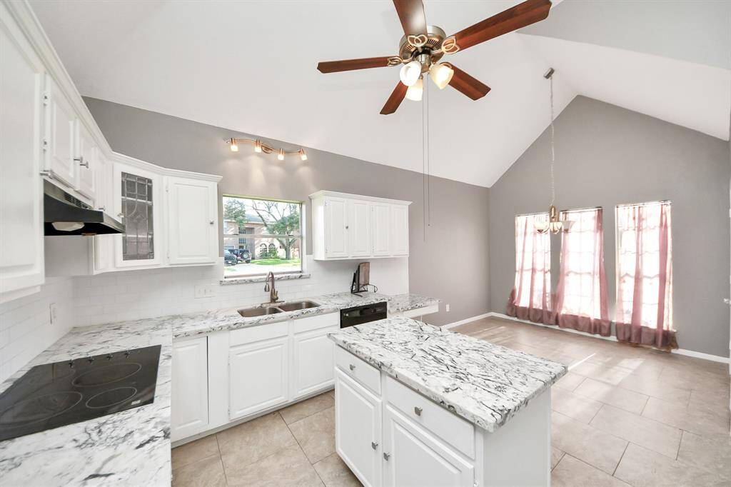 Bright and spacious kitchen with white cabinets, marble countertops, and a central island. The open-plan design features vaulted ceilings, a ceiling fan, and large windows that let in plenty of natural light.