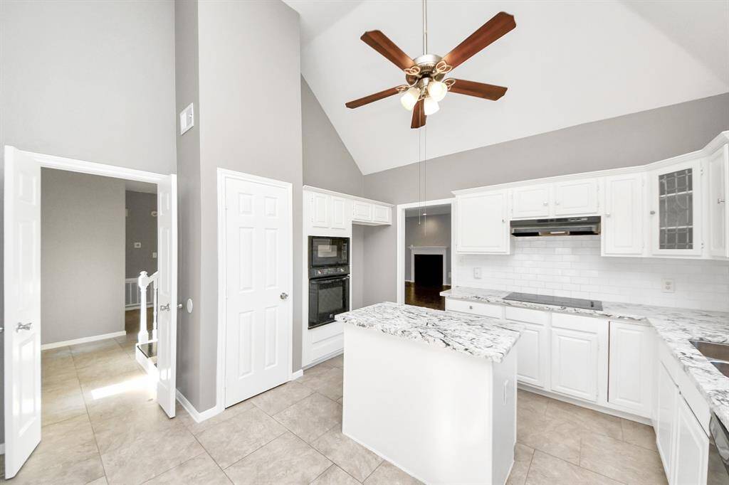 This bright kitchen features vaulted ceilings and a ceiling fan, with modern white cabinetry and granite countertops. It includes a central island, tile flooring, and a double oven, offering a spacious and functional cooking area.