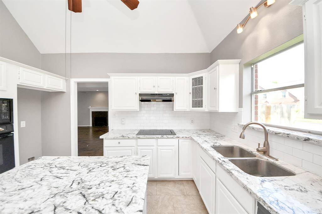 This bright kitchen features high ceilings, granite countertops, white cabinetry, and a large window for natural light. A modern double sink and sleek appliances complete the space, creating a clean and inviting atmosphere.