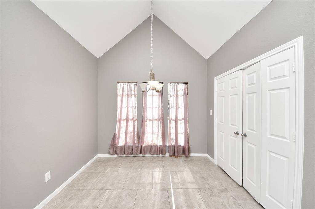 This room features high vaulted ceilings, neutral gray walls, and tiled flooring. It includes a central light fixture, two windows with sheer curtains for natural light, and double doors, offering versatile space for dining or a small office.