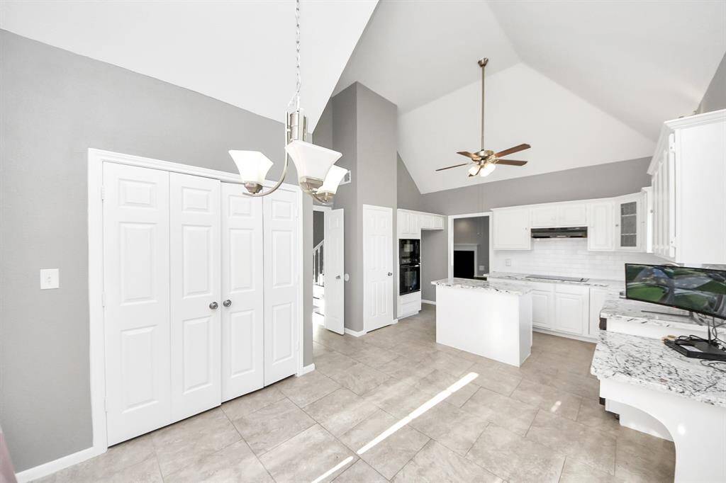 This photo showcases a bright, spacious kitchen with high vaulted ceilings and modern finishes. It features white cabinetry, a central island, and a tiled backsplash. A ceiling fan and stylish light fixture add functionality and elegance, while neutral tones create a welcoming atmosphere.