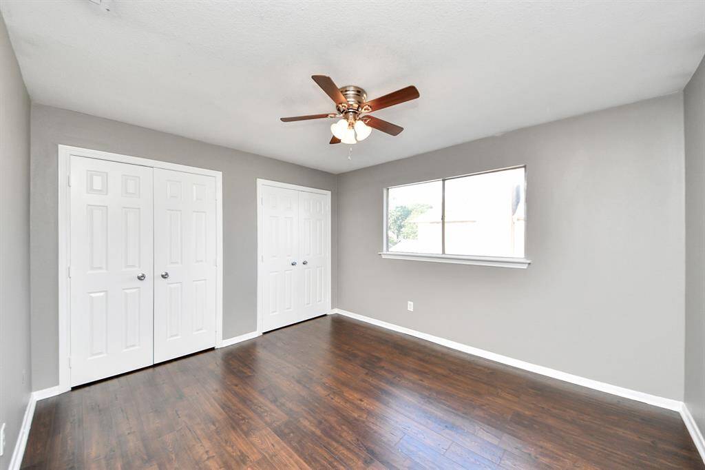 This room features hardwood flooring, neutral gray walls, and a ceiling fan with lights. It has ample natural light from a large window and includes double-door closets for storage.