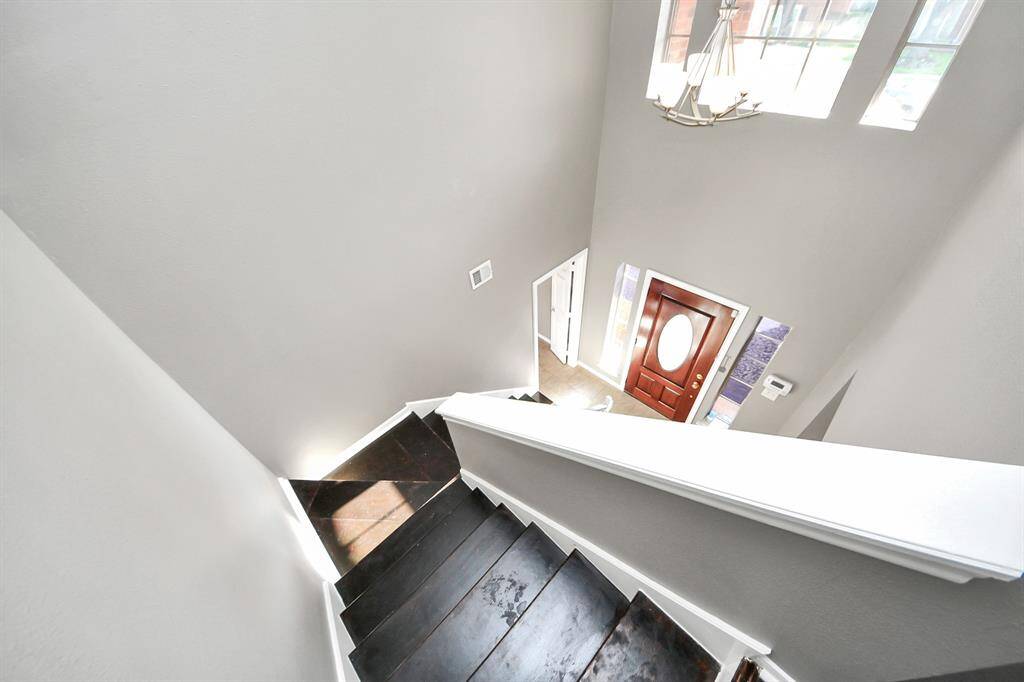 Bright and airy stairway with dark wood steps, leading down to a foyer. Features a stylish wooden front door with an oval glass insert, large windows, and a hanging light fixture, enhancing the open feel.