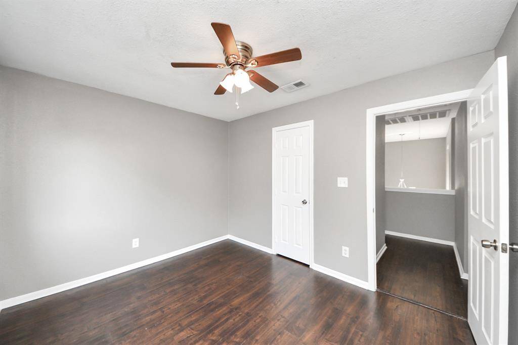 This is a bright, freshly painted room with dark wood flooring, a ceiling fan, and ample natural light. It features a white door and a view into a hallway, offering a modern and clean look.