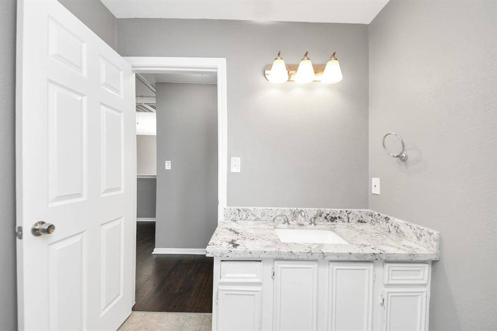 This photo shows a modern bathroom with a granite countertop and white cabinetry. The walls are painted gray, and there's a three-light fixture above the sink. A door opens to an adjacent room with dark wood flooring.